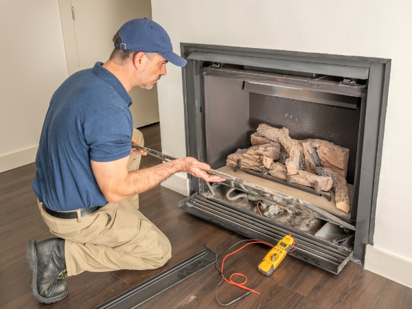 A man inspects a fireplace with tools and a multimeter on the floor beside him.