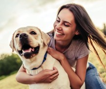 A smiling woman hugs a happy yellow Labrador dog outdoors on a sunny day.