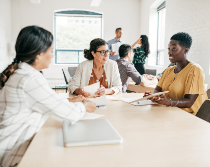 Three women sit at a table having a discussion, while others talk in the background of an office.