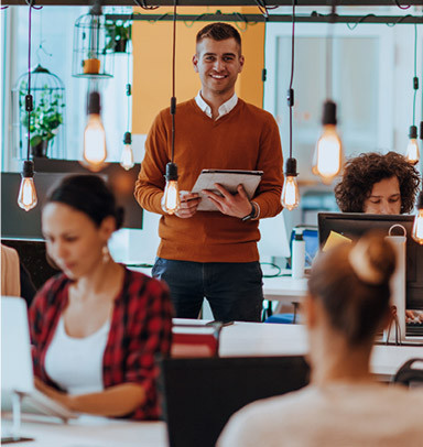 Man holding a tablet stands smiling in a modern, open office with coworkers at desks.