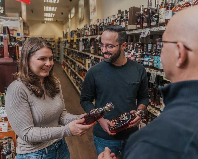 man and woman holding whiskey bottles speaking to store worker
