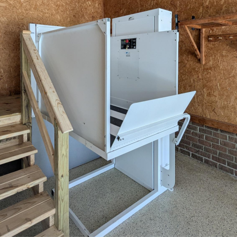 White, metal wheelchair lift next to a wooden staircase inside a room with wood-paneled walls.