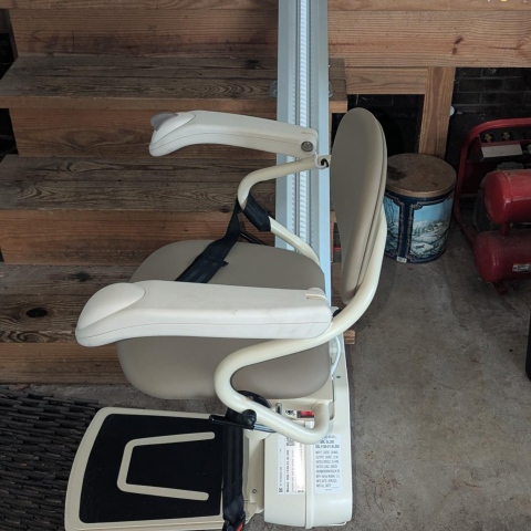 A stairlift chair is positioned at the base of a wooden staircase in a garage or basement.