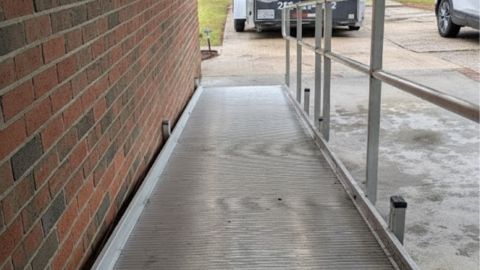 A metal wheelchair ramp runs alongside a brick wall toward a driveway with parked vehicles.