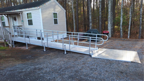 A metal wheelchair ramp leads from a gravel driveway to the entrance of a small house.