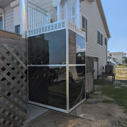 Outdoor residential elevator with glass panels beside a house with light siding and a deck.