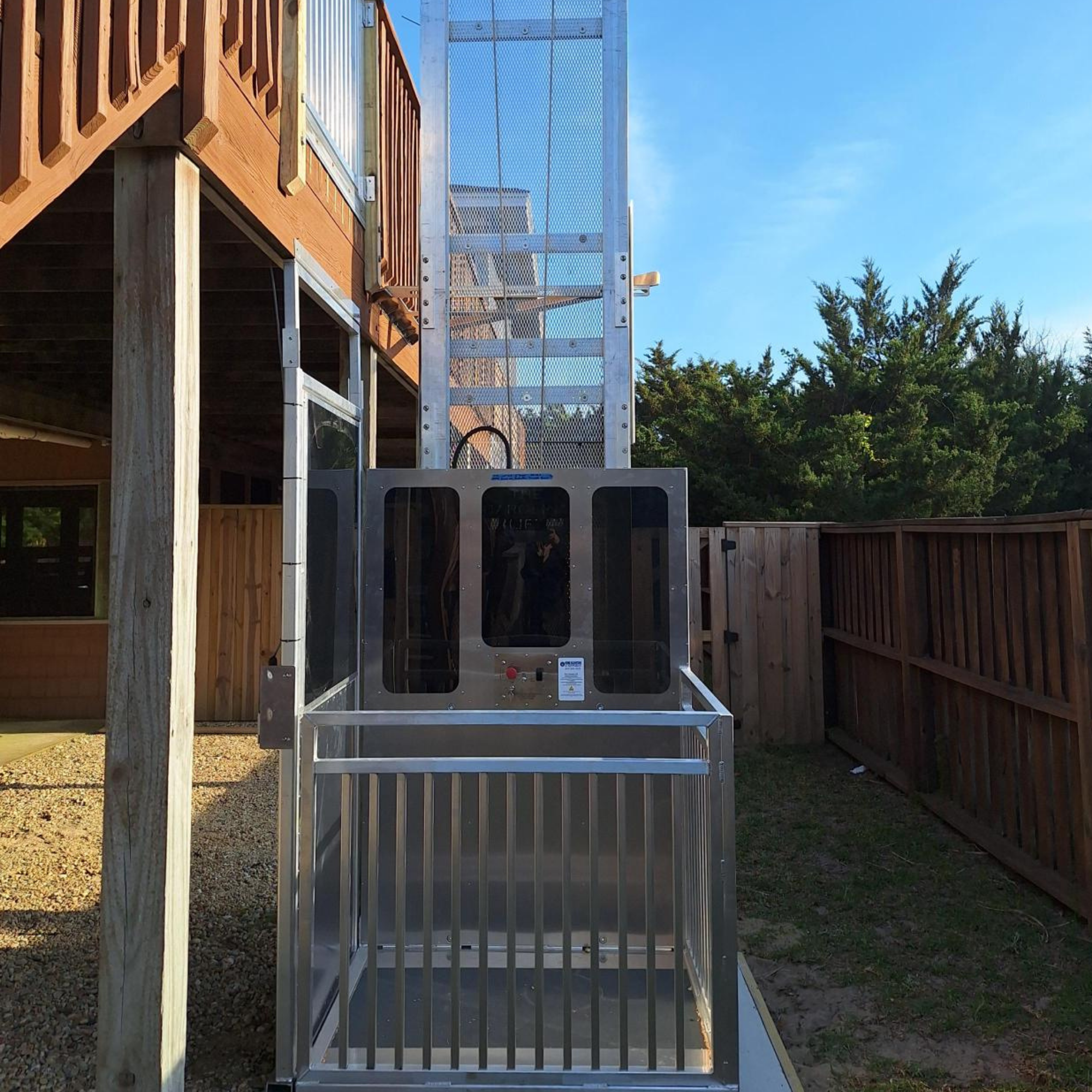 Outdoor residential wheelchair lift by a wooden deck, surrounded by a fenced yard and trees.