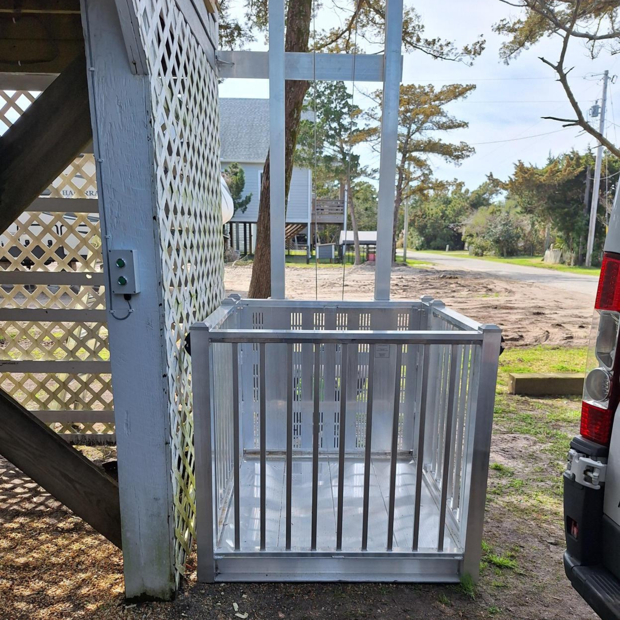 A small outdoor elevator cargo lift with metal railings beside a wooden staircase and a house.
