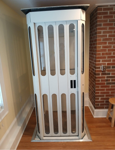 A small white home elevator with glass panels, located indoors near a brick wall and window.