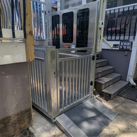An outdoor wheelchair lift beside porch stairs, with a Beware of the Dog sign on the wall.