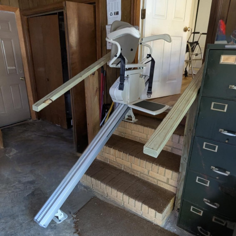 A stair lift with wooden rails installed on brick steps going from a garage to a house interior.
