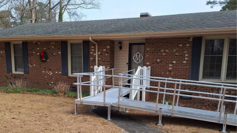 A wheelchair ramp leads to the front door of a brick house with dark shutters and a wreath.