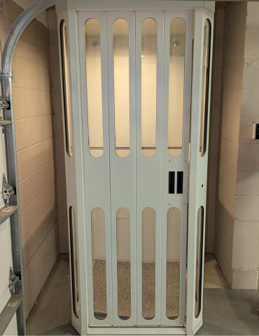 White residential elevator with vertical oval cutouts, located in a basement or garage space.