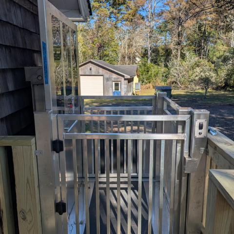A metal wheelchair lift at an outdoor porch, with a yard and shed in the background.