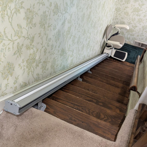 A stairlift installed on a wooden staircase with floral wallpaper on the wall beside it.