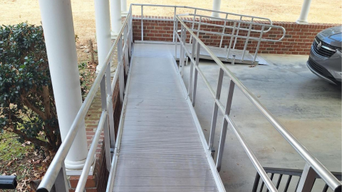 A metal wheelchair ramp with railings leads up to a brick building near a parked car.