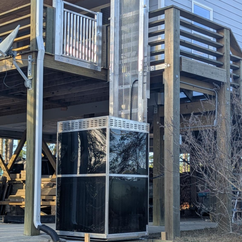 A black outdoor elevator is installed next to a wooden deck and staircase of a house.
