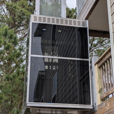 Outdoor residential elevator with glass sides next to a wooden deck, surrounded by trees.