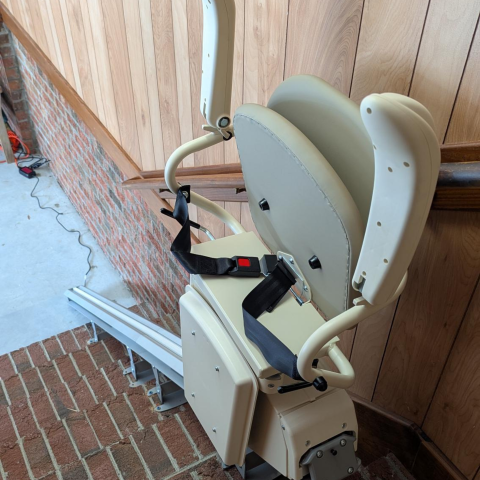 A beige stair lift chair with a seatbelt is installed along a brick staircase and wooden wall.