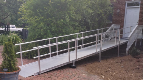 A metal wheelchair ramp leads up to the front door of a brick house with white railings.