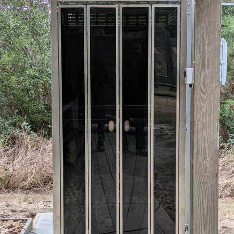 A glass door with metal frames reflecting trees and a wooden pathway outside.