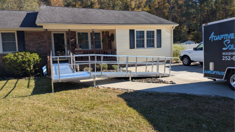 A metal wheelchair ramp leads from a driveway to the front porch of a single-story house.