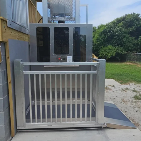 Outdoor wheelchair lift with a metal gate, next to a ramp, by a building and grassy area.