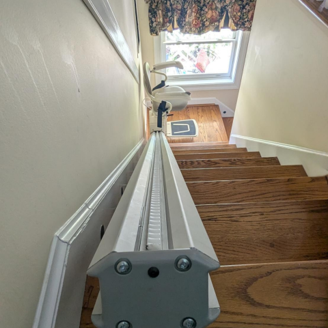 A stair lift runs along a wooden staircase toward a window with floral curtains.