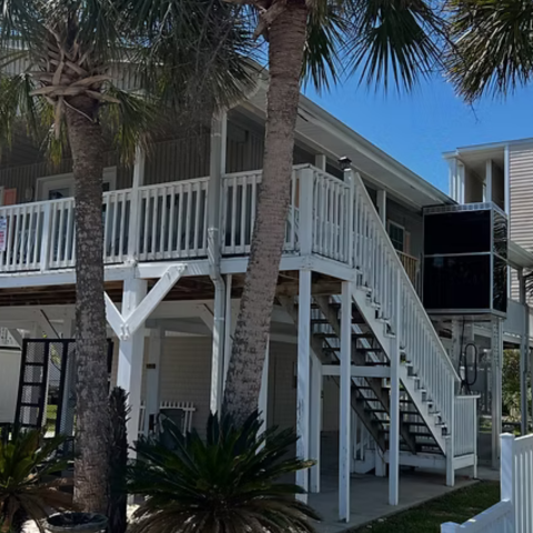 Two-story beach house with white railings, palm trees, and an outdoor staircase under a blue sky.