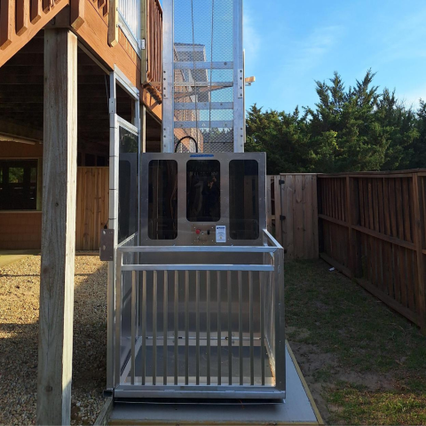 Outdoor residential elevator lift near a wooden deck, surrounded by a fenced yard and trees.