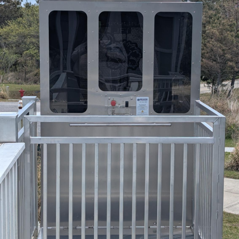 A silver wheelchair lift with glass panels and metal railings outdoors near some trees.