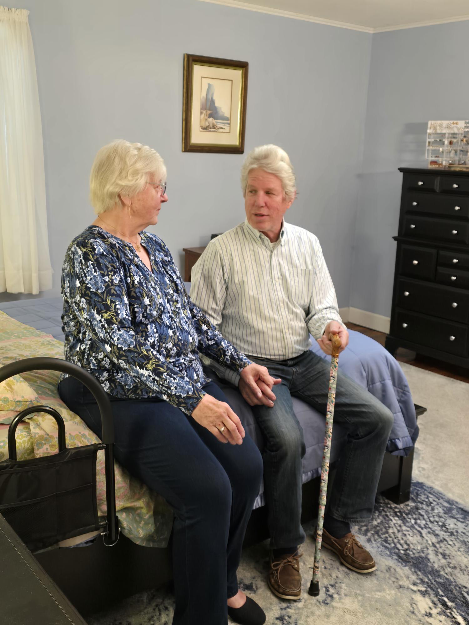 Two older adults sit on a bed in a bedroom, talking; one holds a decorative cane.