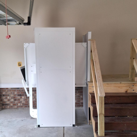 White vertical platform lift installed next to wooden stairs in a residential garage.