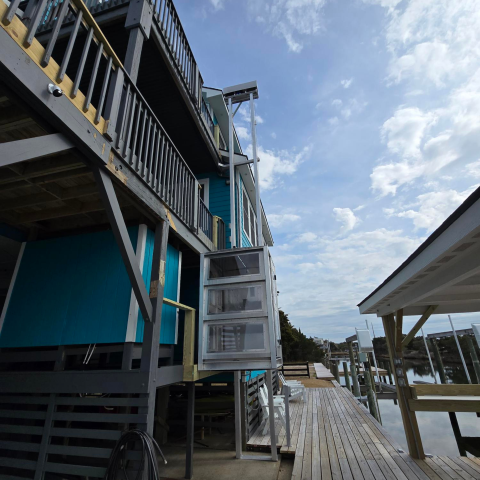 A waterside house with a wooden deck, enclosed platform lift, and partly cloudy sky overhead.
