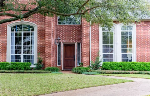 red brick house with large windows, a wooden front door, and green shrubs