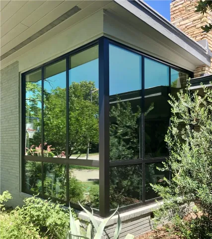 Large corner windows on a modern house reflect trees and greenery under a clear blue sky.