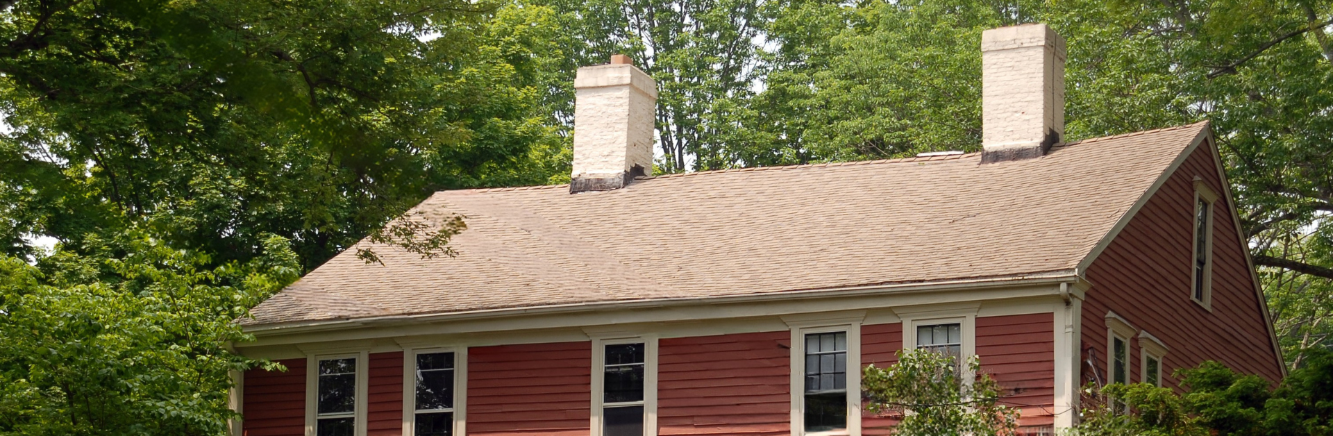 A red house with beige trim and two chimneys, surrounded by green trees and shrubs.