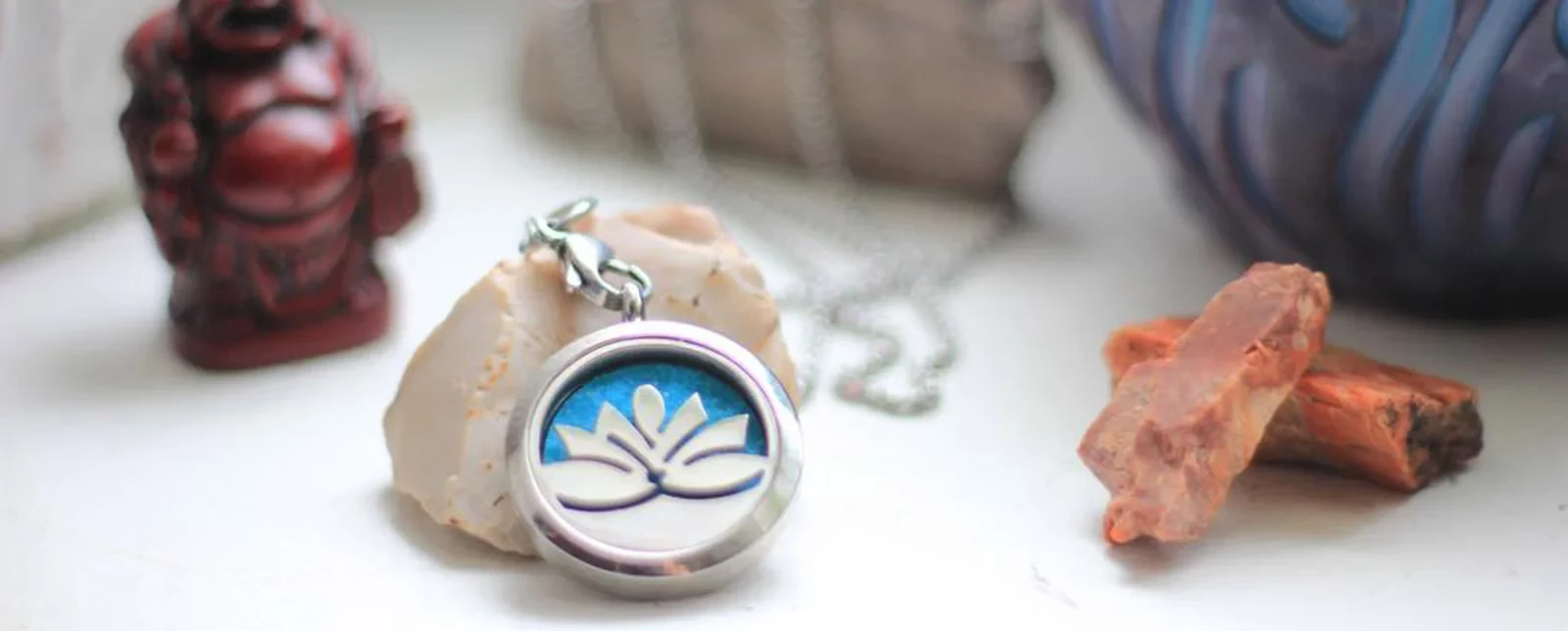 A lotus pendant on a chain rests on a stone, with a Buddha statue and crystal nearby.