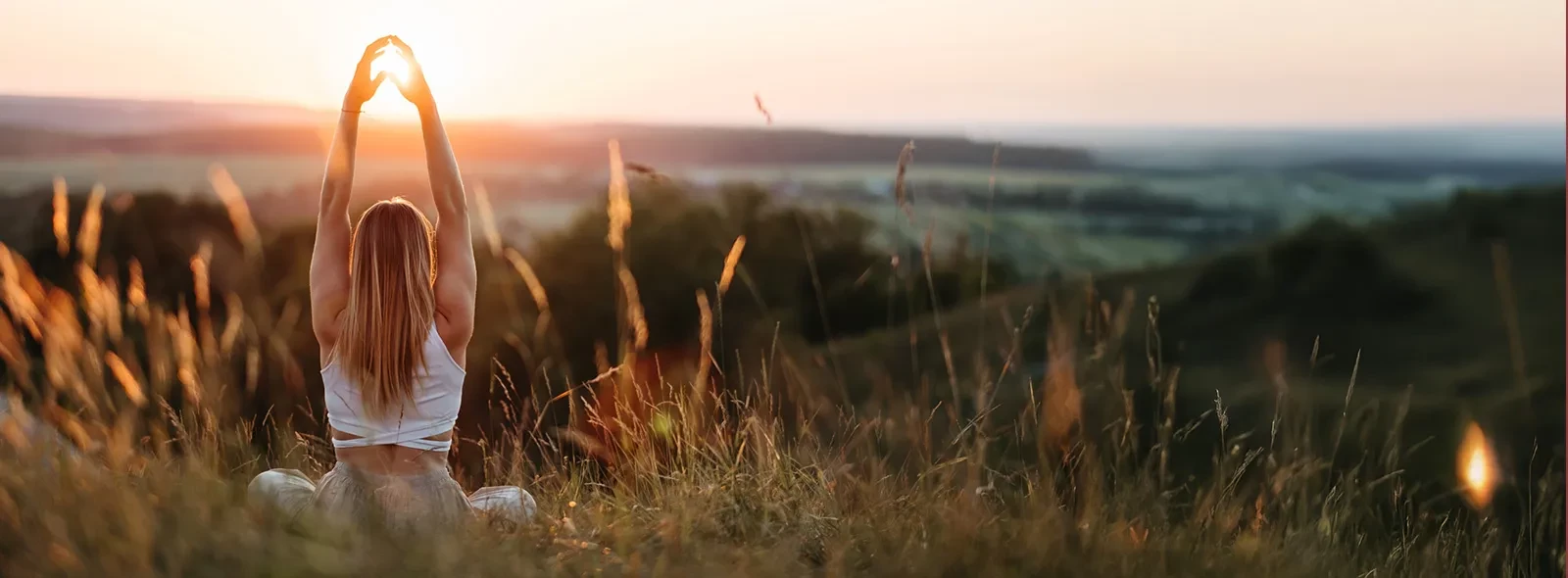 Person sitting in tall grass at sunrise, arms raised overhead, facing a scenic landscape.