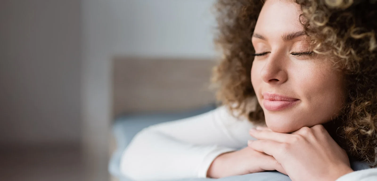 A woman with curly hair smiles peacefully, resting her chin on her hands with eyes closed.