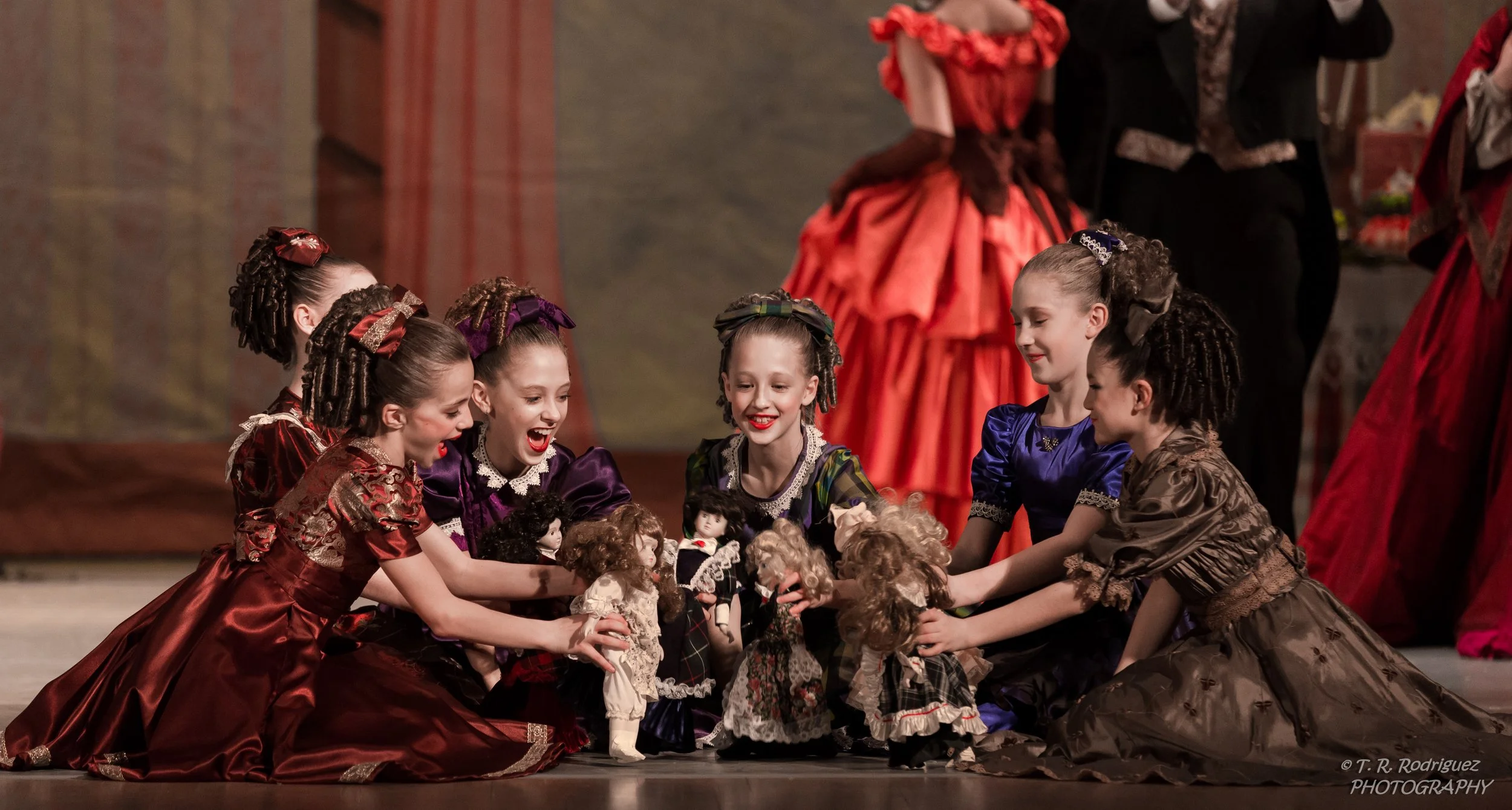 Six young girls dressed in fancy costumes sitting on stage and playing with dolls.