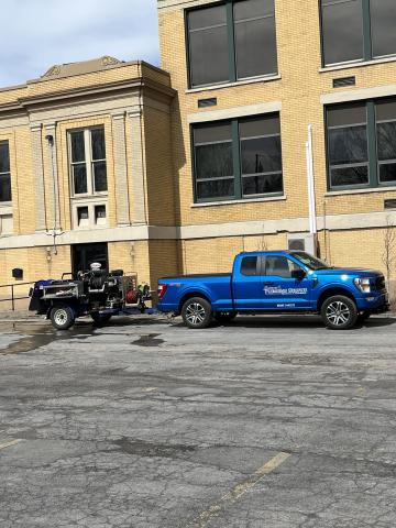 A blue pickup truck with a trailer is parked outside a tan brick building on a sunny day.