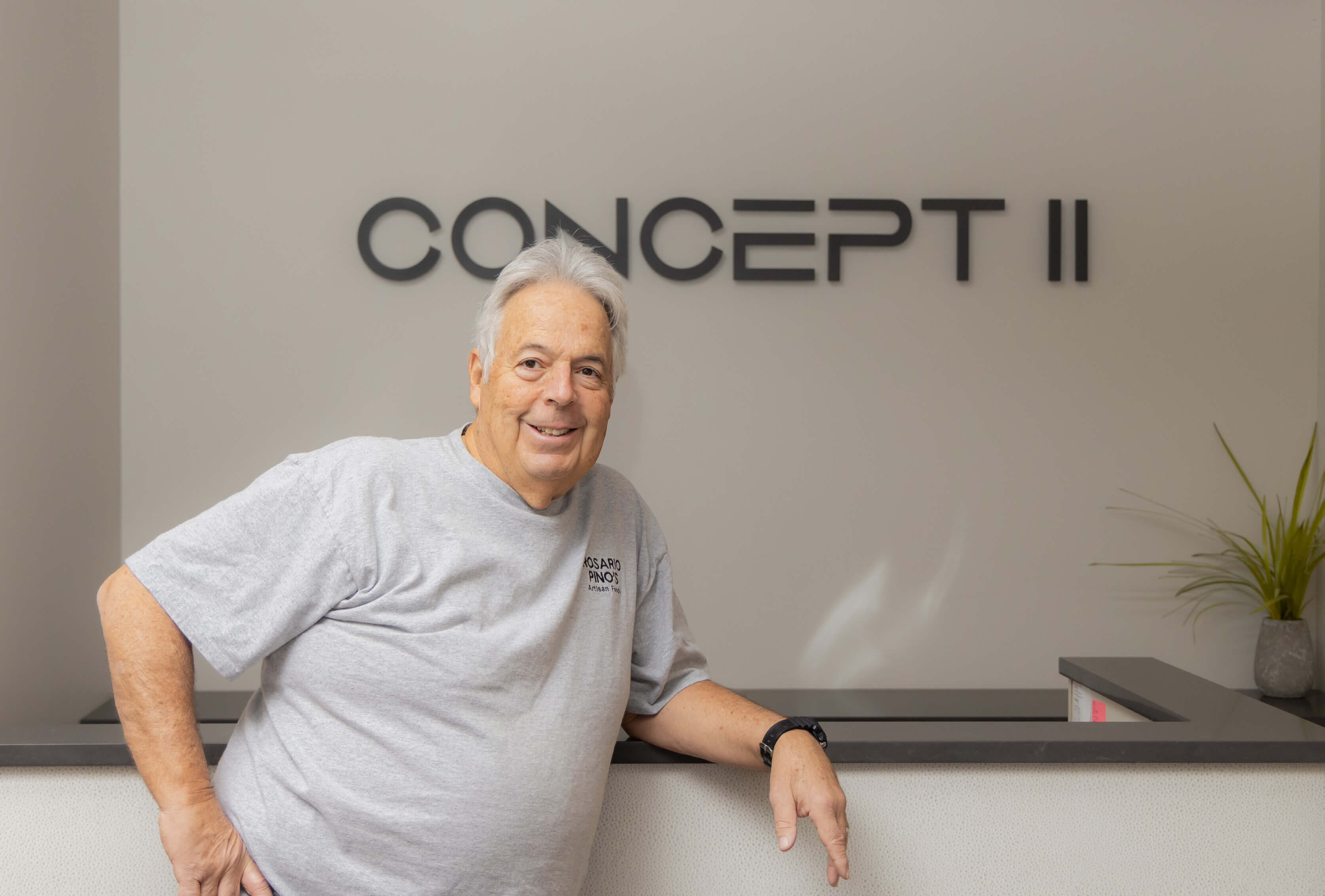 Older man smiling and leaning on a counter with CONCEPT II sign on the wall behind him.