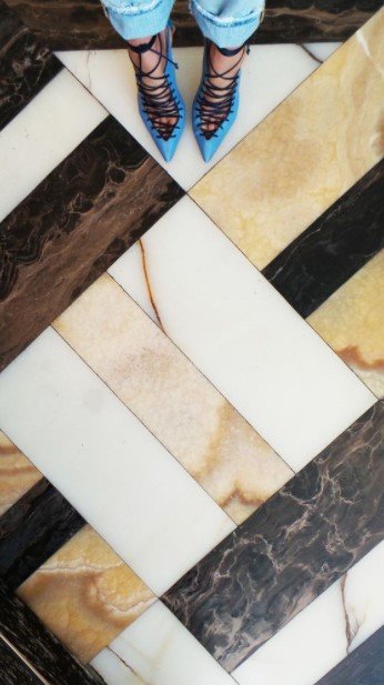 Woman in blue pointed heels and cuffed jeans standing on a patterned marble floor.