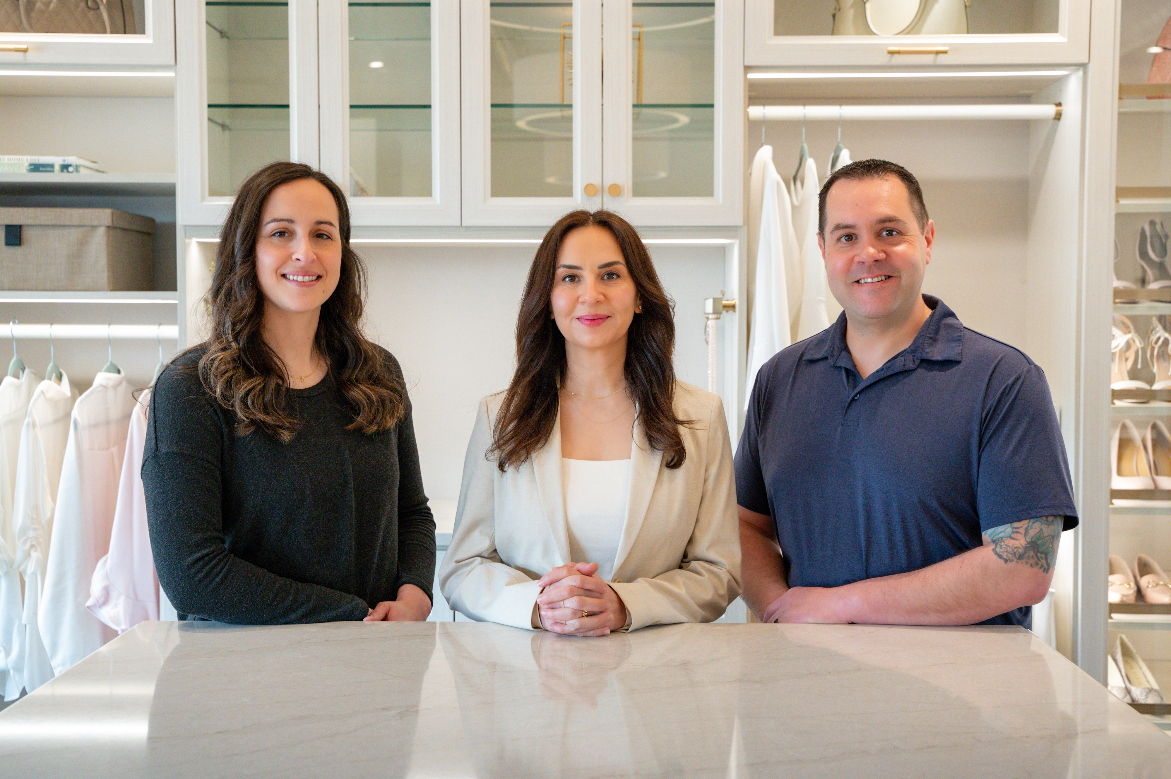 Three people stand side by side, smiling, in a bright, modern walk-in closet.