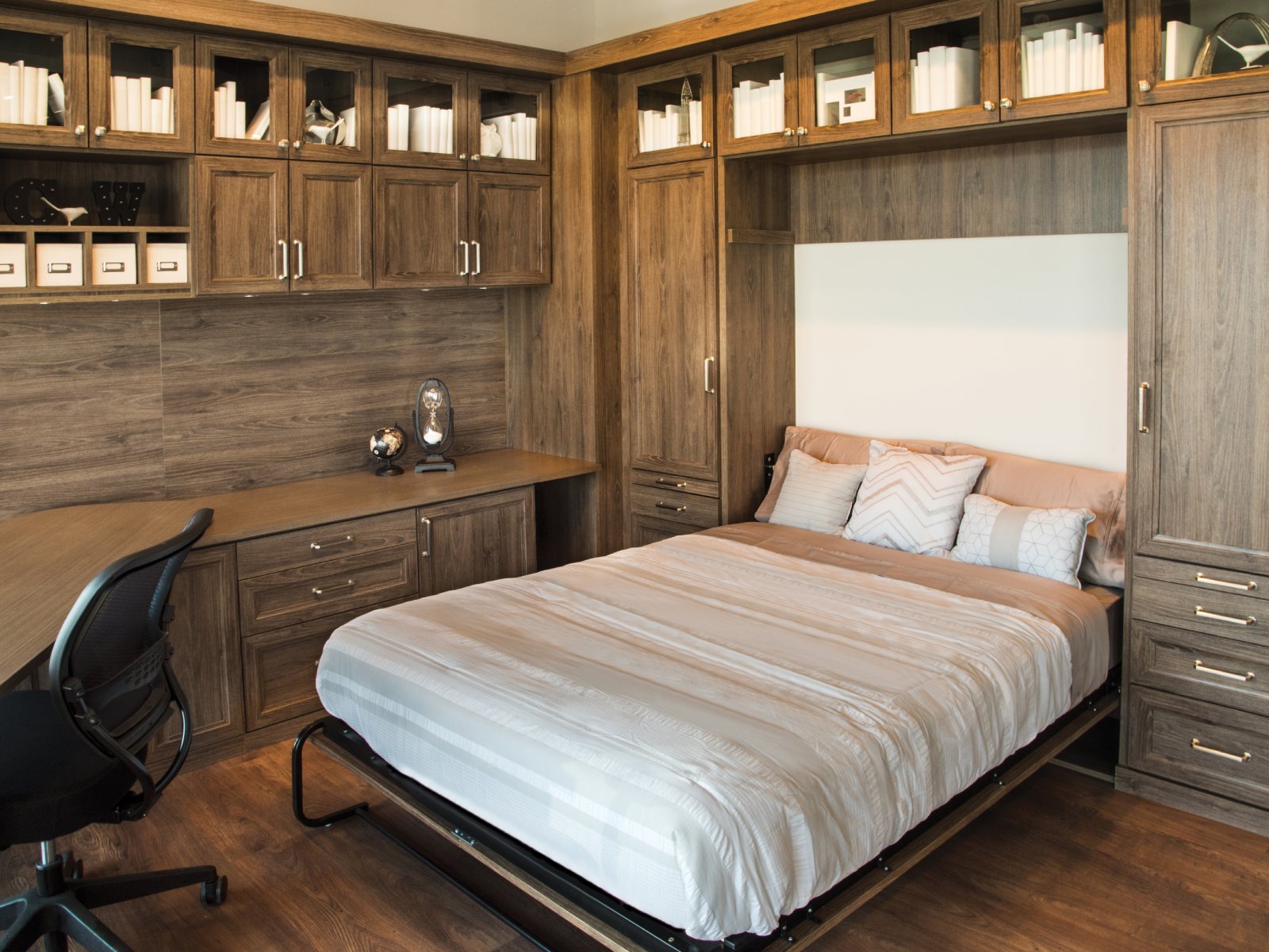 A Murphy bed pulled down in a home office with wooden cabinets and a desk chair.