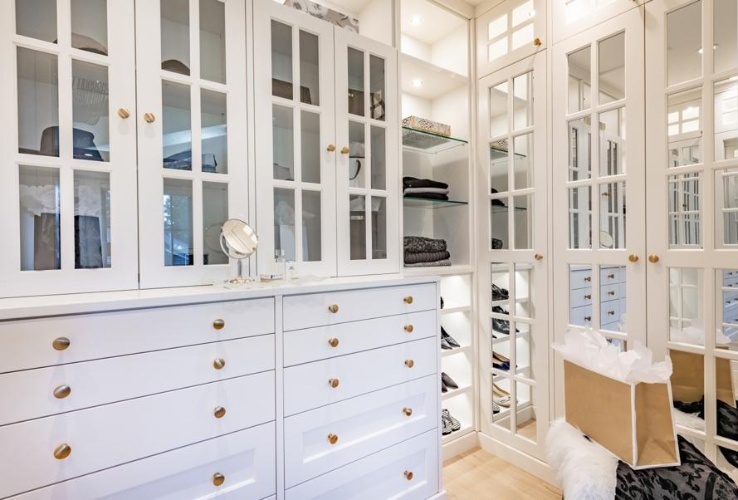 Bright walk-in closet with white cabinets, glass doors, shelves, and a shopping bag on the floor.