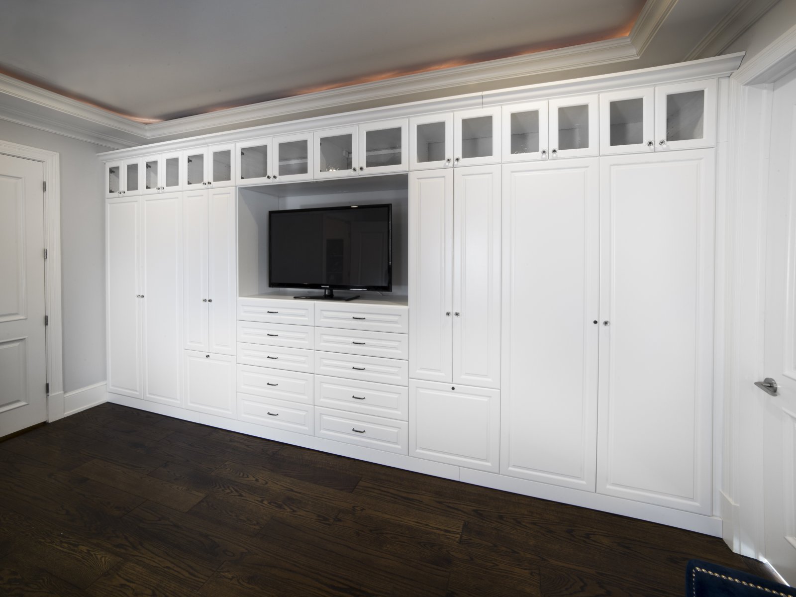Wall of white built-in cabinets with drawers and a TV in the center, on dark wood floor.