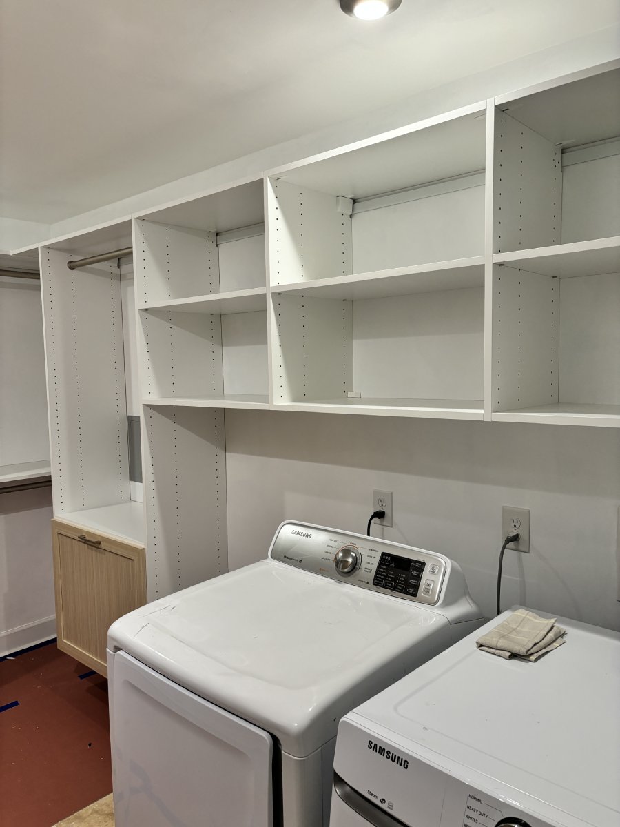 White laundry room with washer, dryer, open shelves, and a small wooden cabinet on the left.