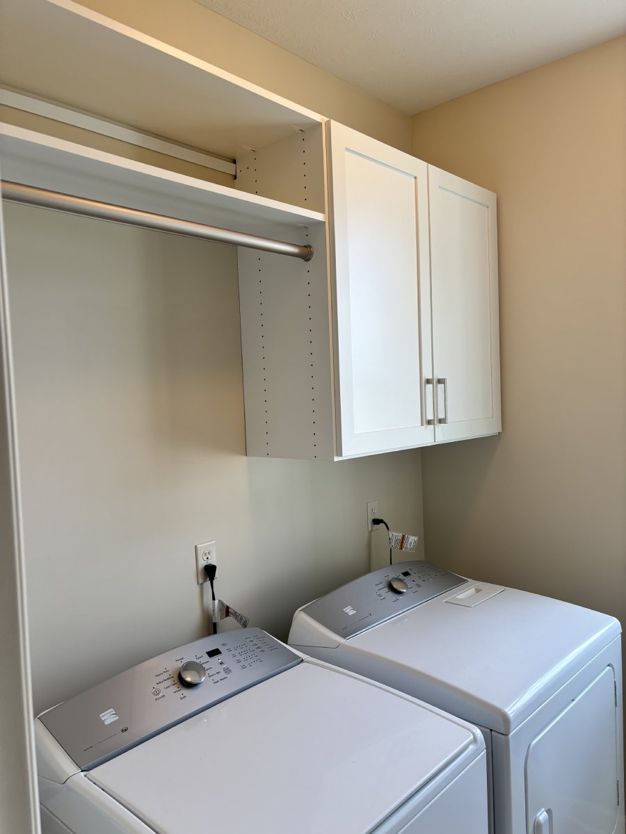 A laundry room with a washer, dryer, white cabinet, and empty open shelving above.